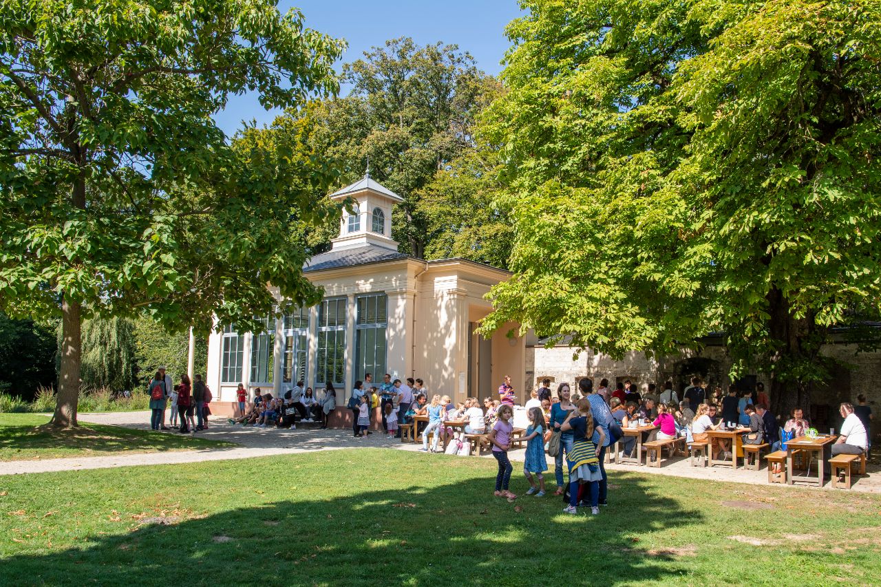 Menschen in der Sonne, die im Sarasinpark Riehen miteinander plaudern. Kinder spielen im Vordergrund.